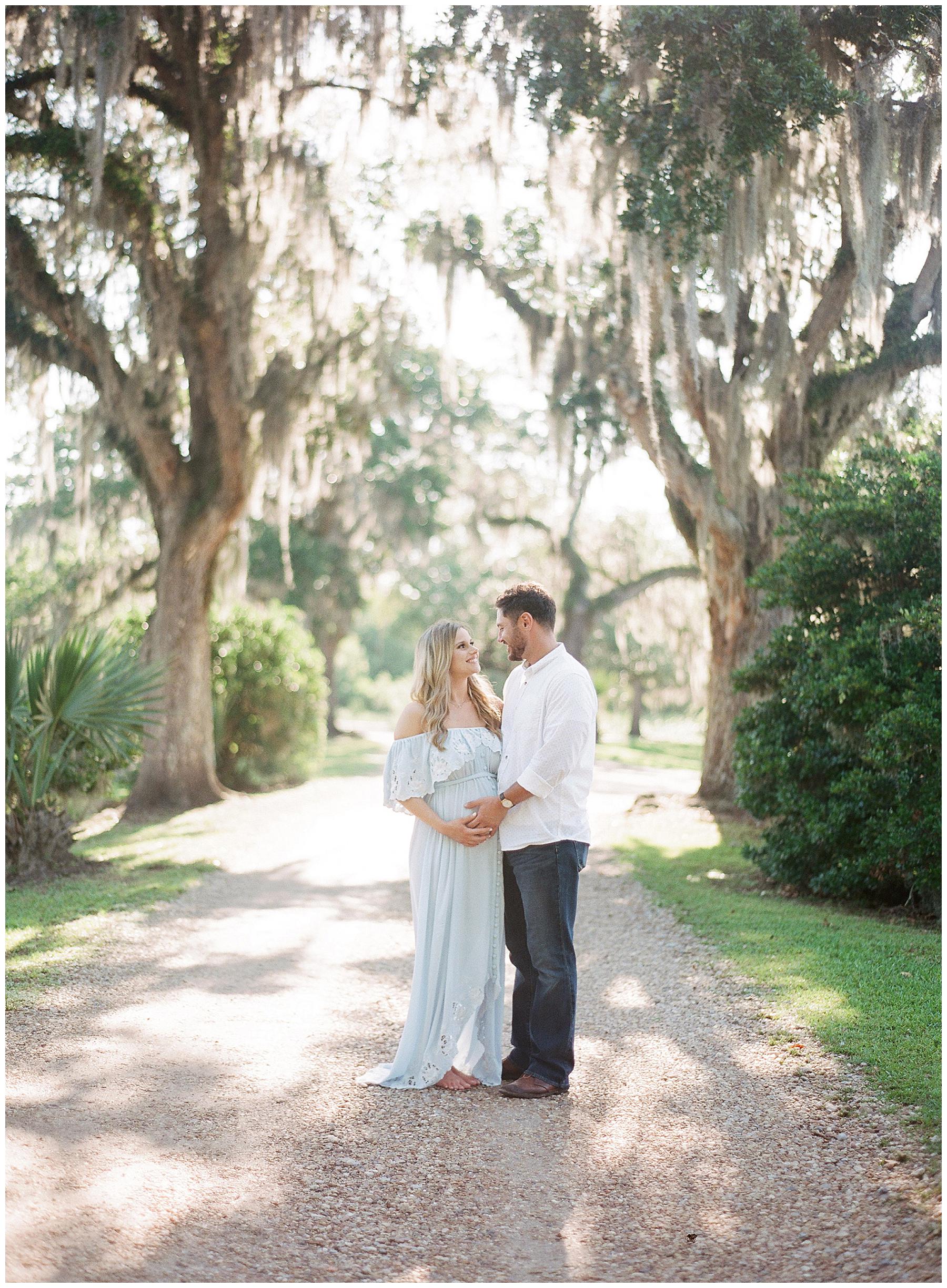 Maternity couple poses for a portrait down gravel road in the louisiana landcape of dripping moss trees