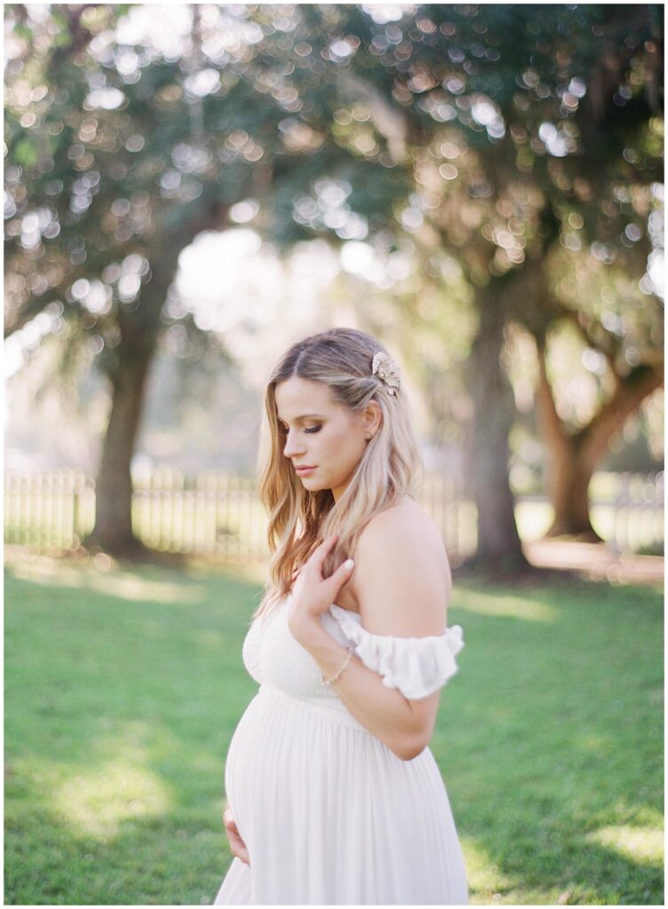 First time mother to be poses in a side profile holding her baby bump with the lush green louisiana landscape as her backdround