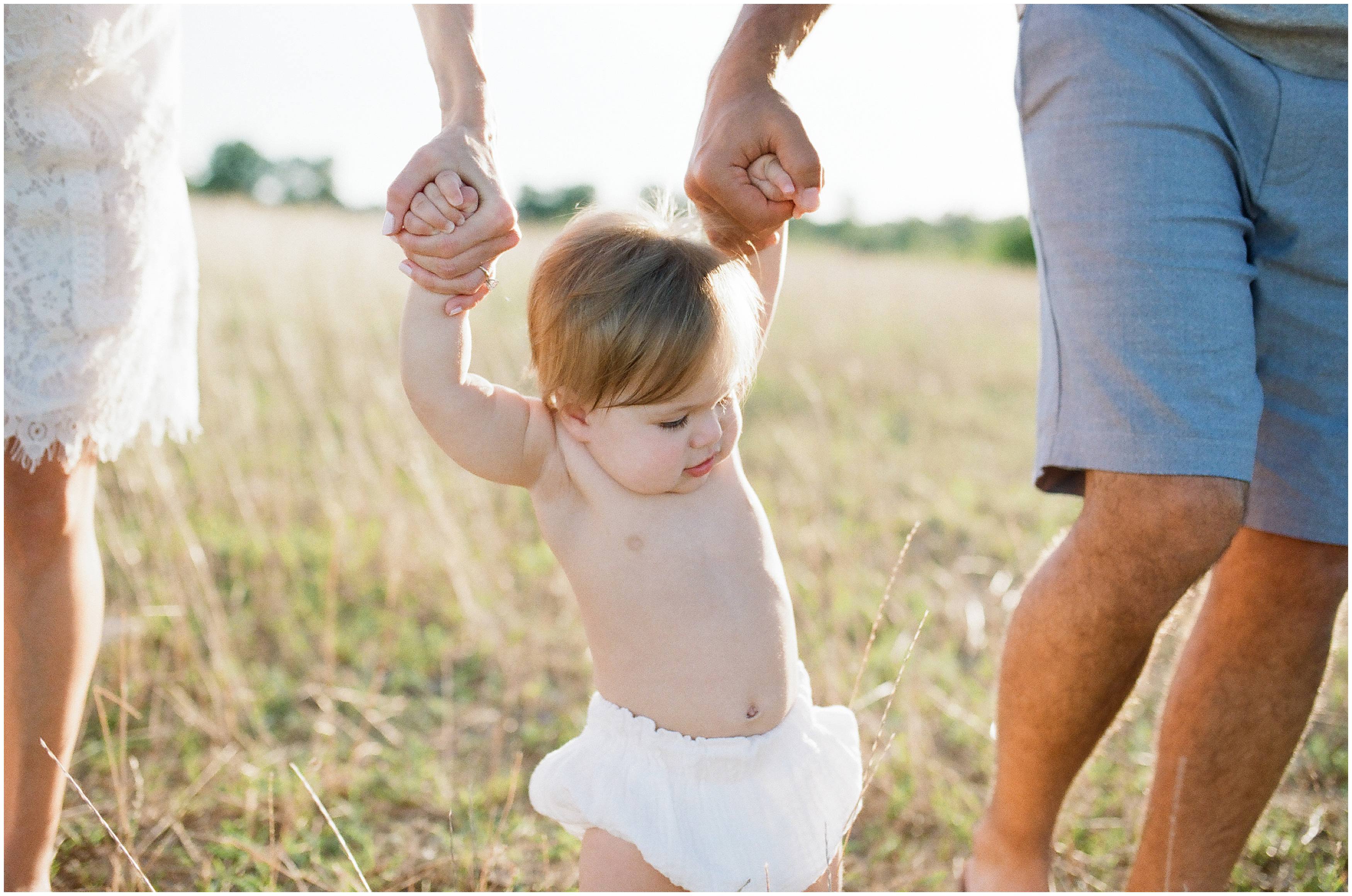 shirtless baby swings between parents arms in a field captured by photographer natural light contax 645