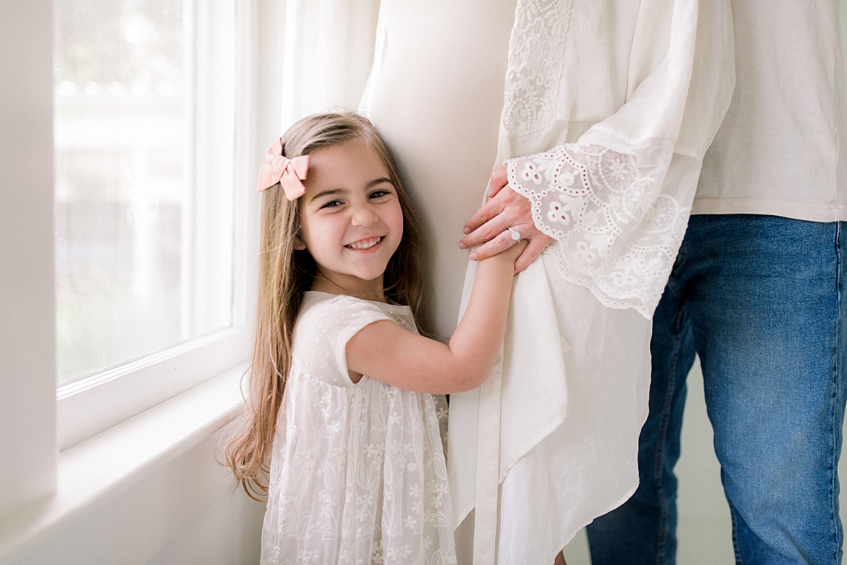 4 year old girl smiles at the camera as she hugs her moms pregnant belly