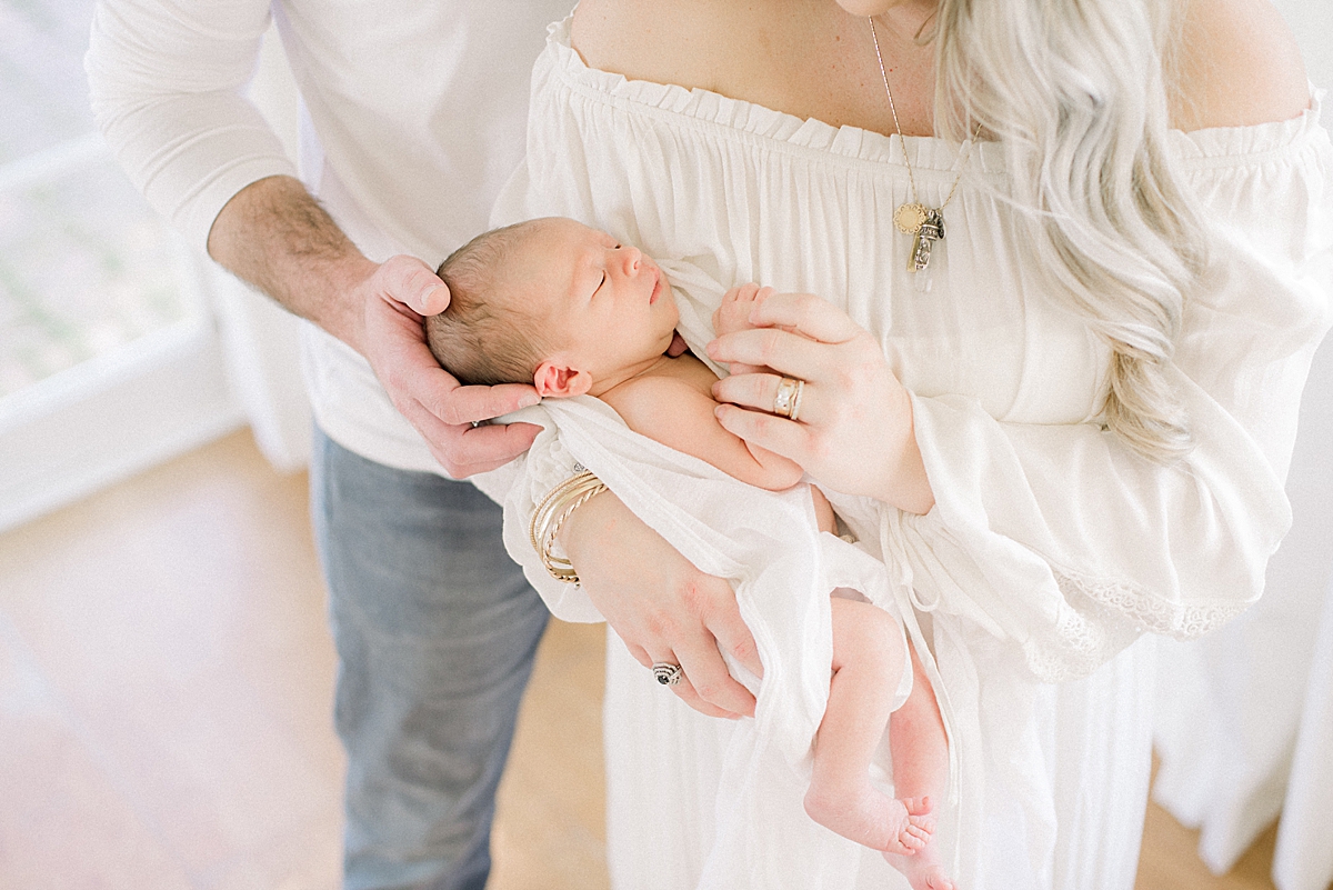 baby sleeps in mothers arms and his little toes dangle down, his dad softly rubs his head and sings a lullaby to get him relaxed 