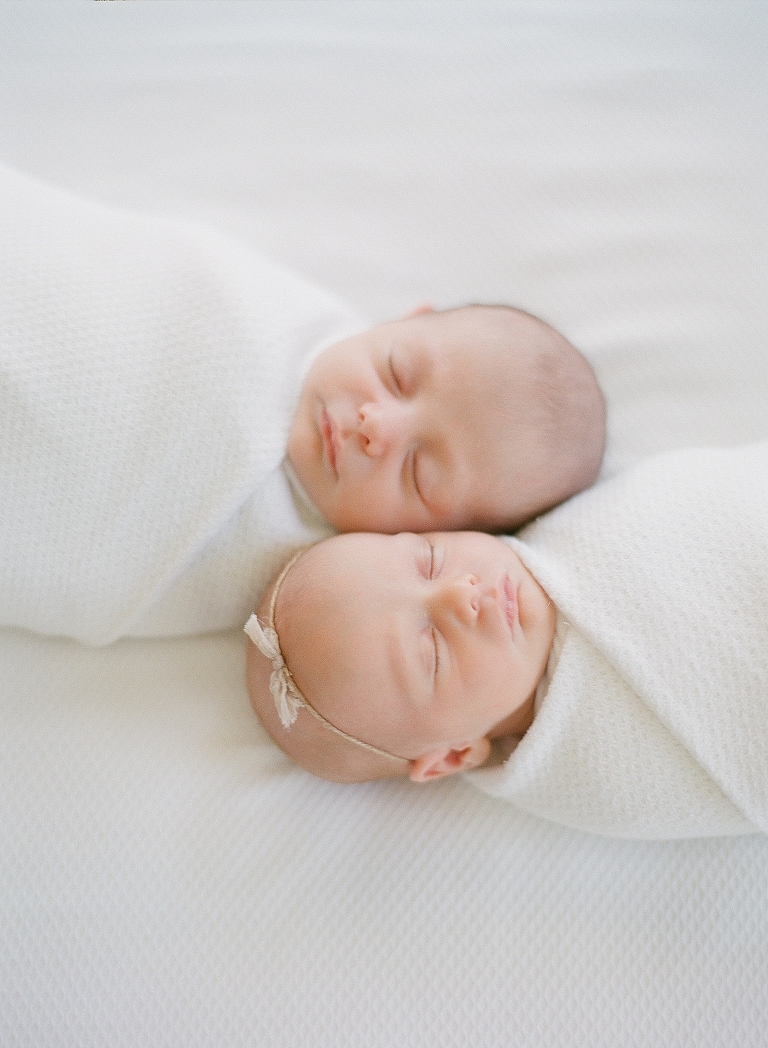 Twin babies swaddles on bed sleeping together Girl has on tieback newborn headband with bow
