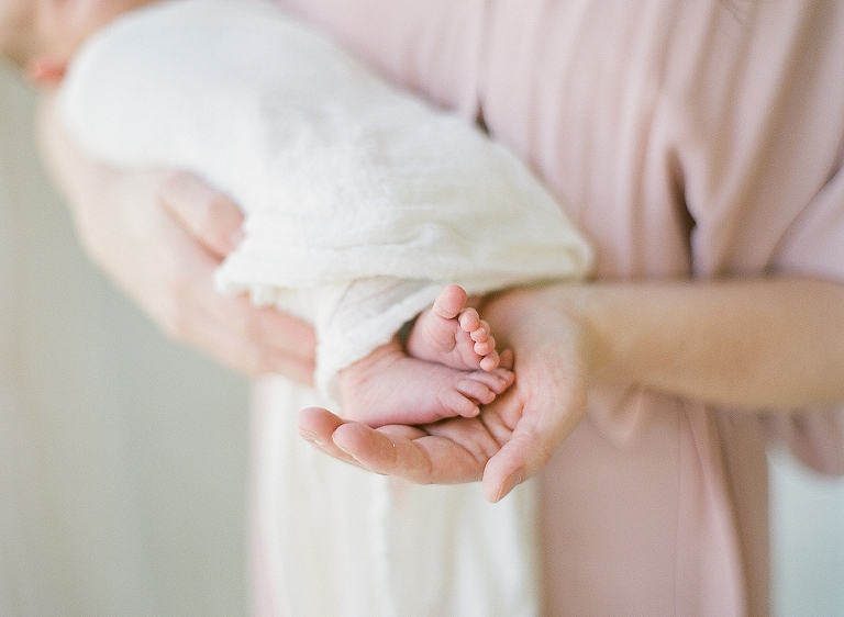 mom holds her newborn baby swaddled in white muslin blanket babies feet are cradled in her hands so soft and sweet