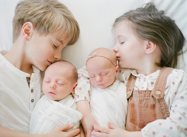 big brother and big sister softly kiss the heads of newborn twins during their photography session 