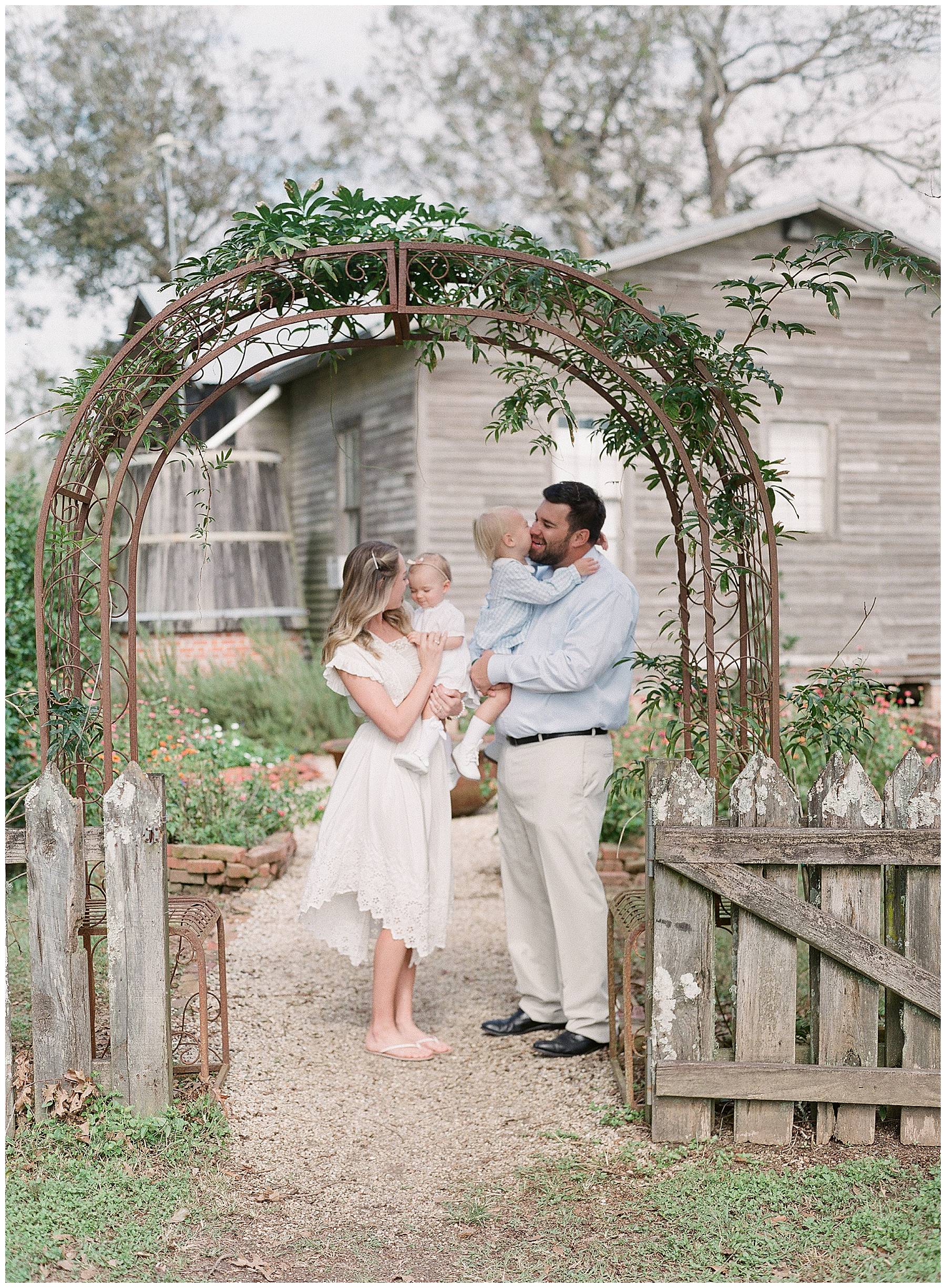 Mom and dad in a butterfly garden holding their babies and kissing them as they giggle. 