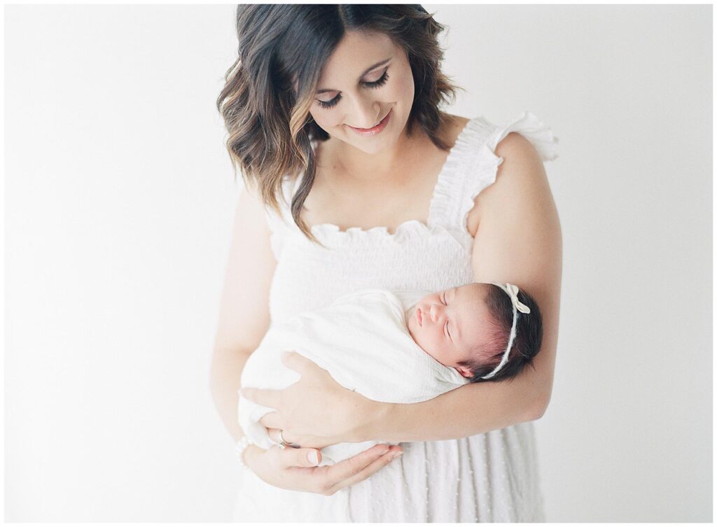 Mother holding newborn baby girl swaddled in white wrap in photographers studio